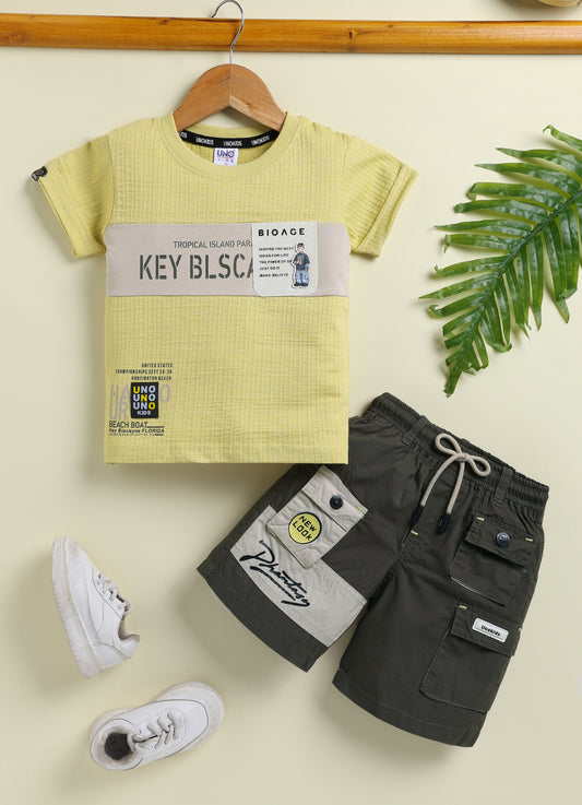 Image file 10407-Lemon.jpg showing a flat lay of a Unokids brand boys two-piece clothing set featuring a textured lemon yellow short-sleeve t-shirt with urban typographic graphics and dark olive green cargo shorts with an elastic drawstring waist and multiple functional pockets.