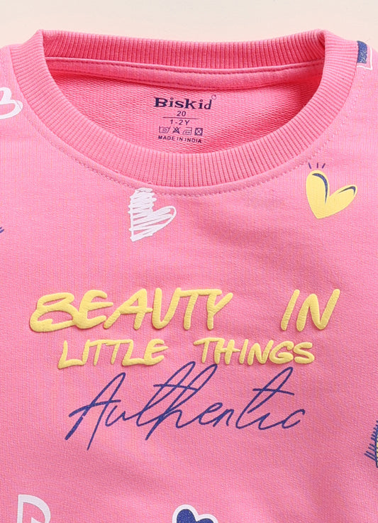 Image file 1751-Pink.jpg showing a Biskid brand girls pink sweatshirt featuring an all-over heart print and the text "BEAUTY IN LITTLE THINGS" with "Authentic" on the front, displayed on a wooden hanger with children's white sneakers and a decorative toy camera nearby.