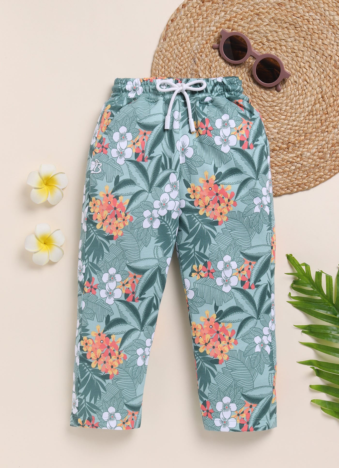 Image file 1511-M.Green.jpg showing a pair of Biskid brand girls mint green joggers with an all-over tropical floral print, featuring an elastic waistband with a white drawstring, displayed with decorative sunglasses, flowers, and greenery.
