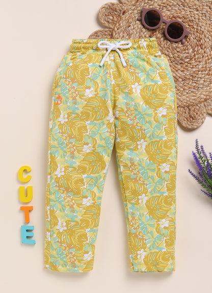 Image file 1511-Mustard.jpg showing a pair of Biskid brand girls mustard yellow joggers with an all-over tropical leaf print, displayed with fun decorative accents like sunglasses and wooden "CUTE" letters.