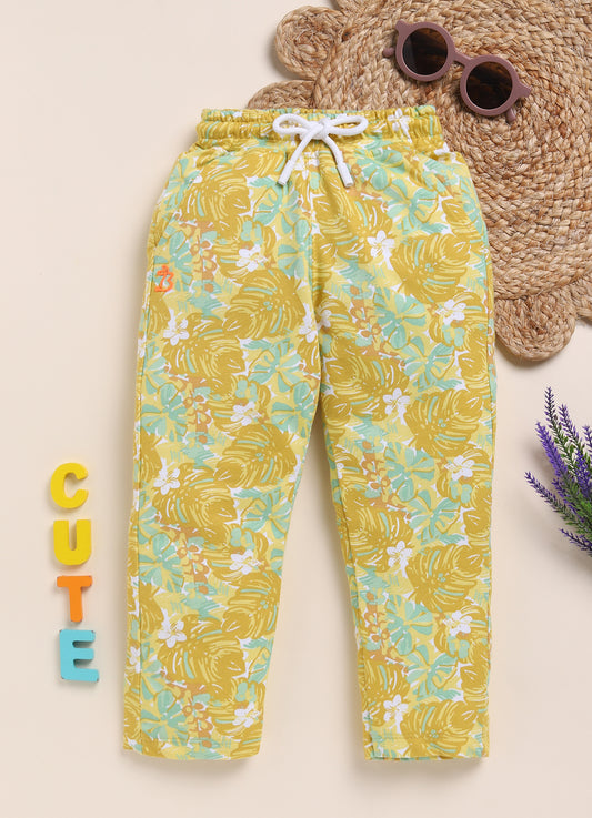 Image file 1511-Mustard.jpg showing a pair of Biskid brand girls mustard yellow joggers with an all-over tropical leaf print, displayed with fun decorative accents like sunglasses and wooden "CUTE" letters.