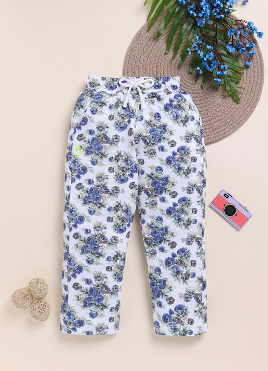 Image file 1511-Navy.jpg showing a pair of Biskid brand girls white joggers with an all-over floral rose print in navy and gray, displayed with a woven mat, blue floral accents, and a toy camera decoration.