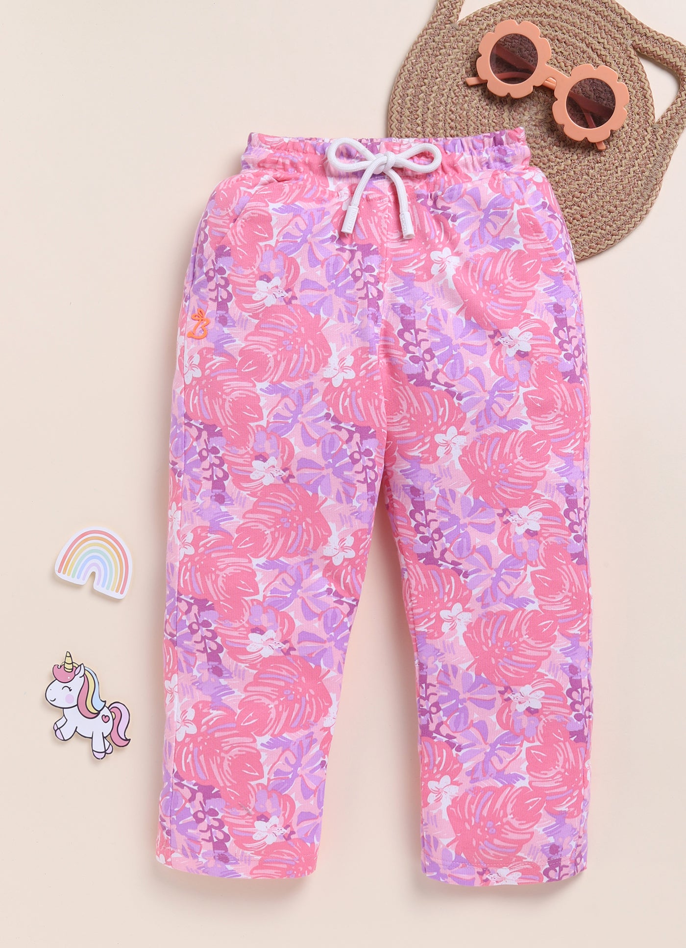 Image file 1511-Pink.jpg showing a pair of Biskid brand girls pink joggers with an all-over tropical leaf print, displayed with fun decorative accents like a rainbow, a unicorn sticker, and flower-shaped sunglasses.