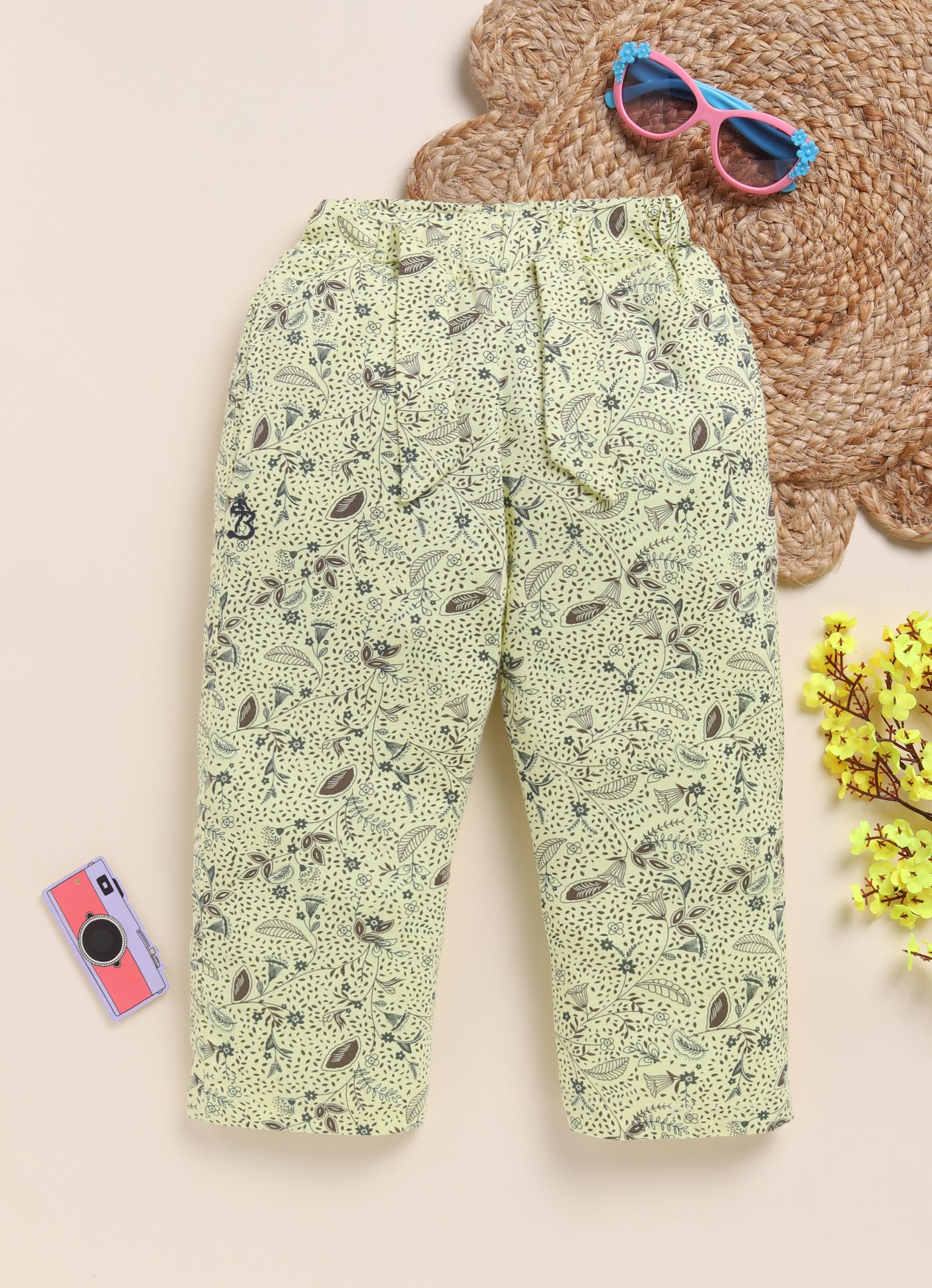 Image file 1512-Lemon.jpg showing a pair of Biskid brand girls' lemon yellow long pants with an all-over botanical print, displayed on a woven mat with pink sunglasses, a toy camera, and yellow decorative flowers nearby.
