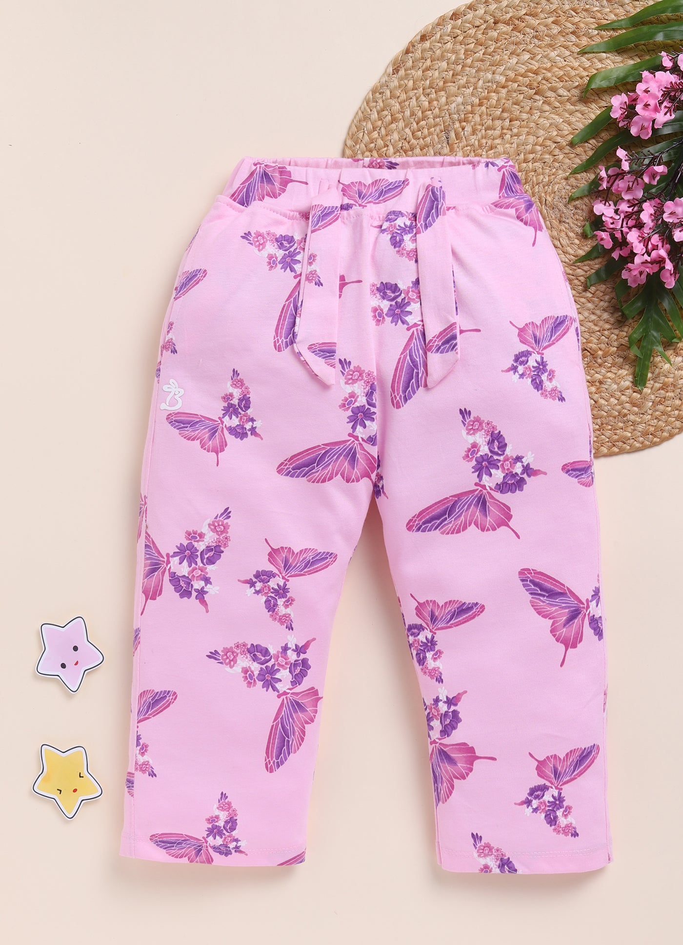 Image file 1512-Pink.jpg showing a pair of Biskid brand girls' pink long pants with an all-over butterfly and floral print, displayed with two star-shaped decorations and a cluster of pink flowers on a woven mat.