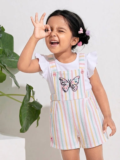 A young girl making a playful face and posing in an Annie brand two-piece clothing set featuring a white flutter-sleeve top under textured short overalls with pastel pink, yellow, and light blue vertical stripes and a large floral butterfly patch on the chest.