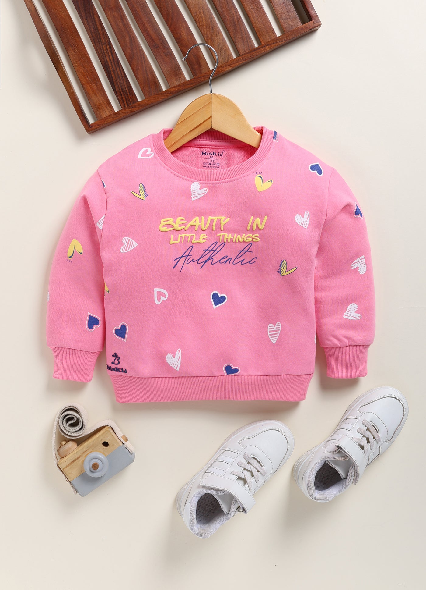 Image file 1751-Pink.jpg showing a Biskid brand girls pink sweatshirt featuring an all-over heart print and the text "BEAUTY IN LITTLE THINGS" with "Authentic" on the front, displayed on a wooden hanger with children's white sneakers and a decorative toy camera nearby.