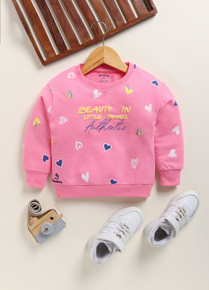 Image file 1751-Pink.jpg showing a Biskid brand girls pink sweatshirt featuring an all-over heart print and the text "BEAUTY IN LITTLE THINGS" with "Authentic" on the front, displayed on a wooden hanger with children's white sneakers and a decorative toy camera nearby.