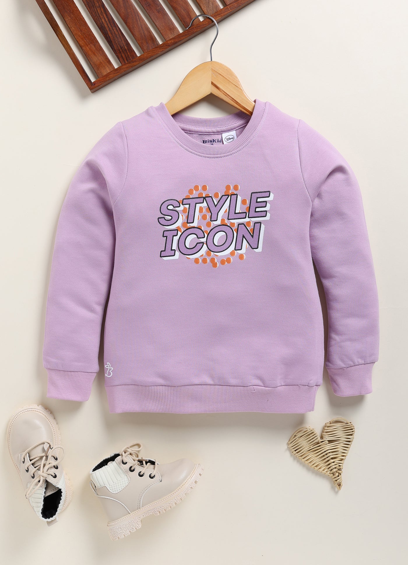 Image file 1754-Purple.jpg showing a Biskid brand girls lavender-purple sweatshirt featuring the text "STYLE ICON" on the front, displayed on a wooden hanger with children's boots and a heart-shaped woven decoration nearby.
