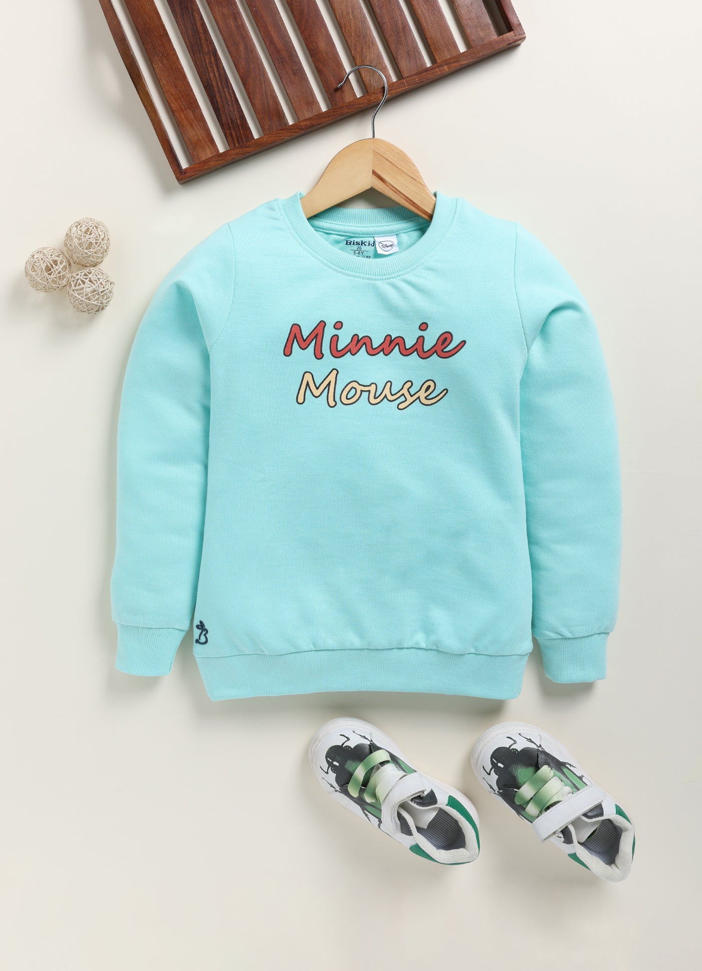 Image file 1754-Skyblue.jpg showing a Biskid brand girls sky blue fleece sweatshirt with "Minnie Mouse" lettering on the front, displayed on a wooden hanger with children's sneakers and decorative spheres nearby.