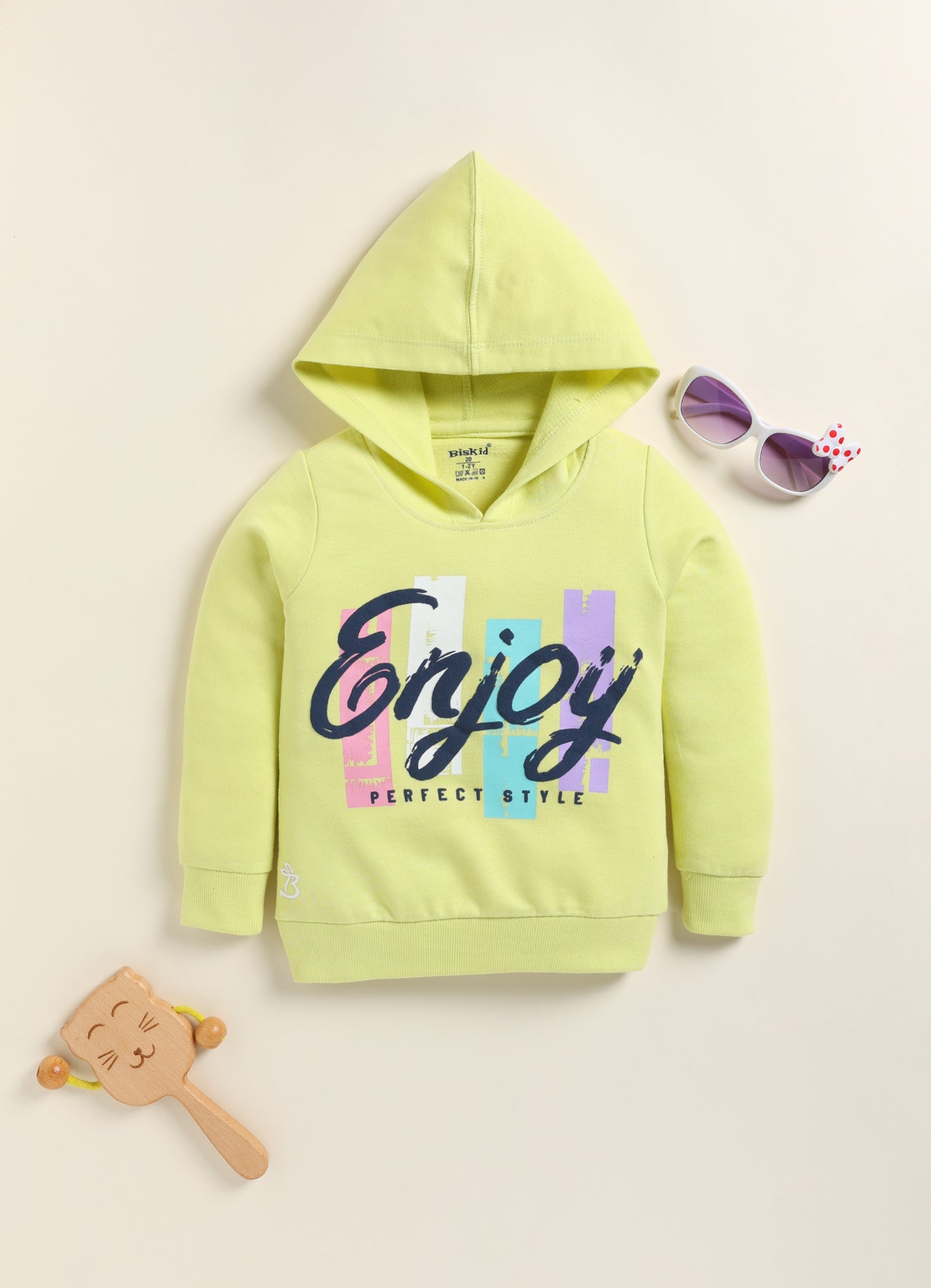 Image file 1756-Green.jpg showing a flat lay of a lime green Biskid brand girls hooded sweatshirt featuring the "Enjoy" graphic and "PERFECT STYLE" text, displayed with decorative items including children's sunglasses and a wooden toy.