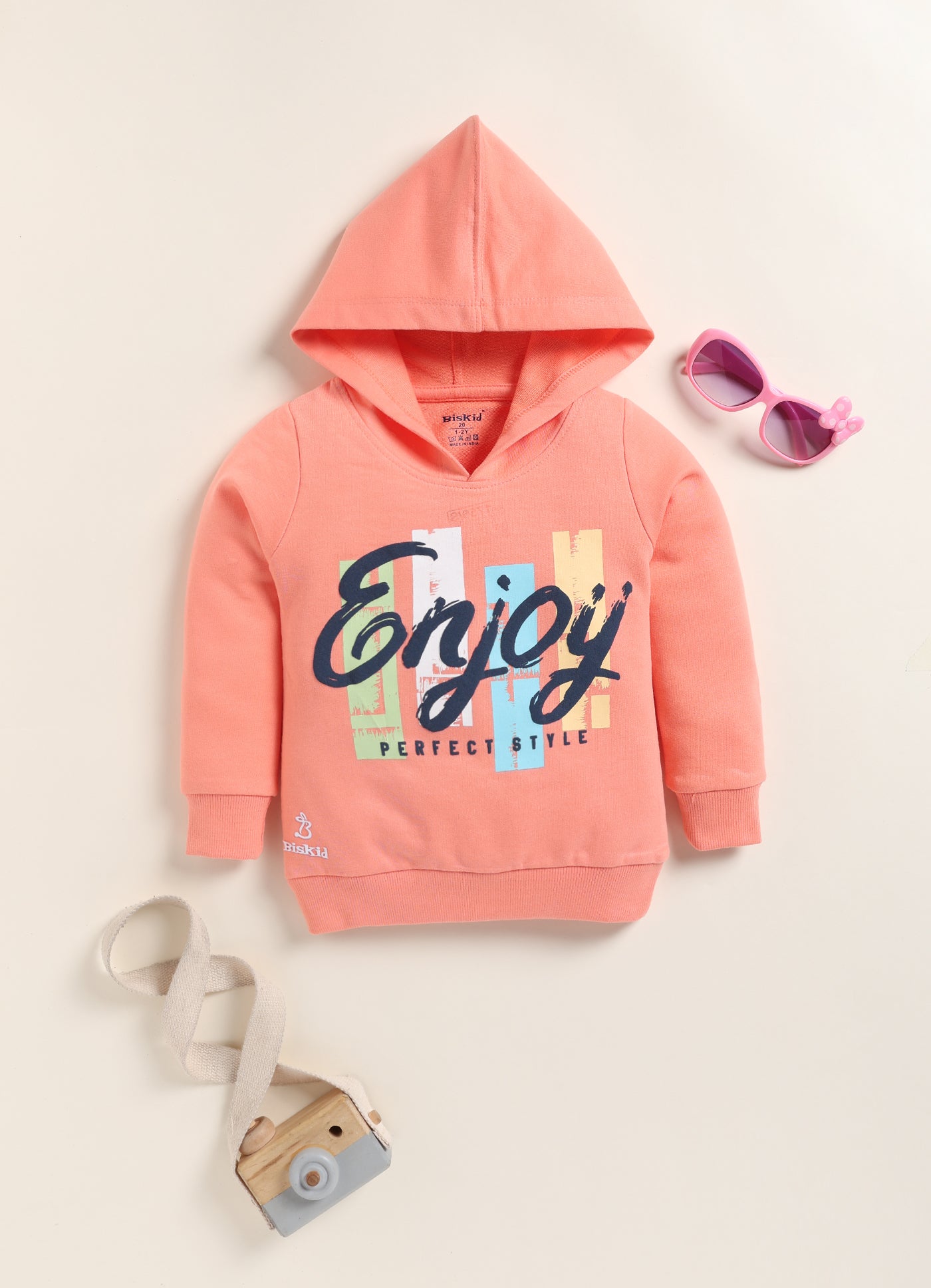 Image file 1756-Peach.jpg showing a flat lay of a peach Biskid brand girls hooded sweatshirt featuring the "Enjoy" graphic and "PERFECT STYLE" text, displayed with decorative items including children's sunglasses and a toy camera.