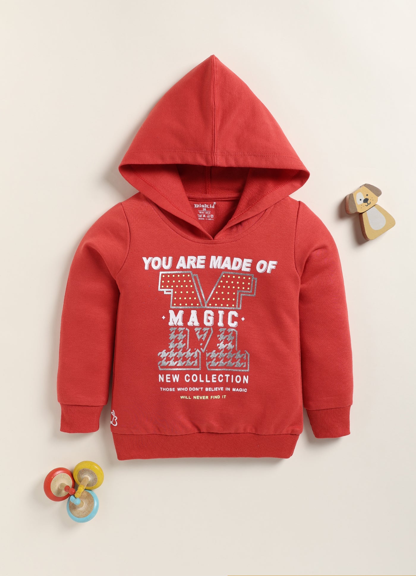 Image file 1756-Red.jpg showing a flat lay of a red Biskid brand girls hooded sweatshirt featuring the text "YOU ARE MADE OF MAGIC" and associated graphic, displayed with decorative items including a small wooden dog toy and colorful spinning tops.
