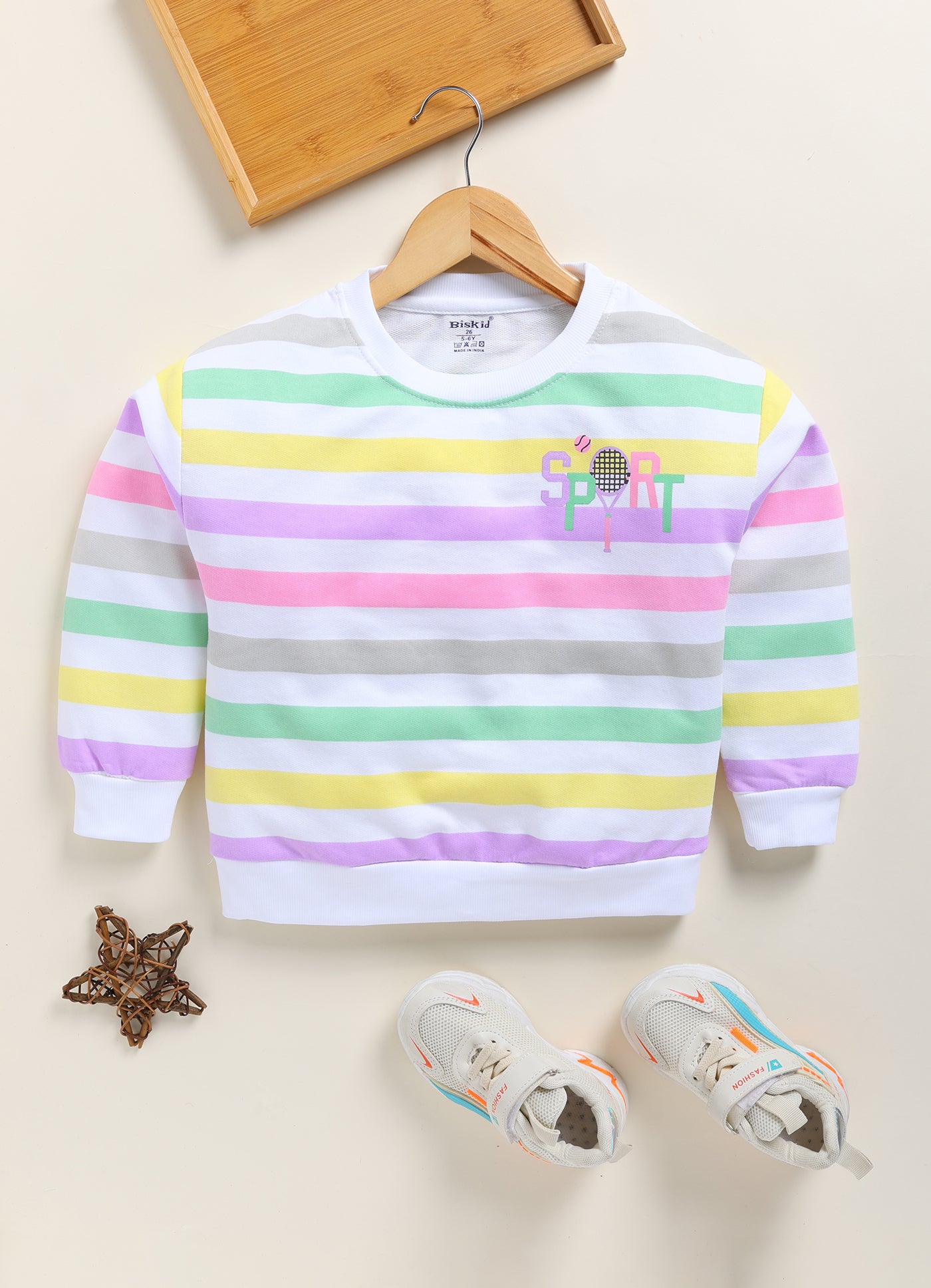 showing a flat lay of a Biskid brand girls long-sleeve pullover top. The top features horizontal stripes in pastel pink, yellow, green, and grey on a white base, with a "SPORT" graphic featuring a tennis racket on the upper chest, and white ribbed trim at the neck, cuffs, and hem.