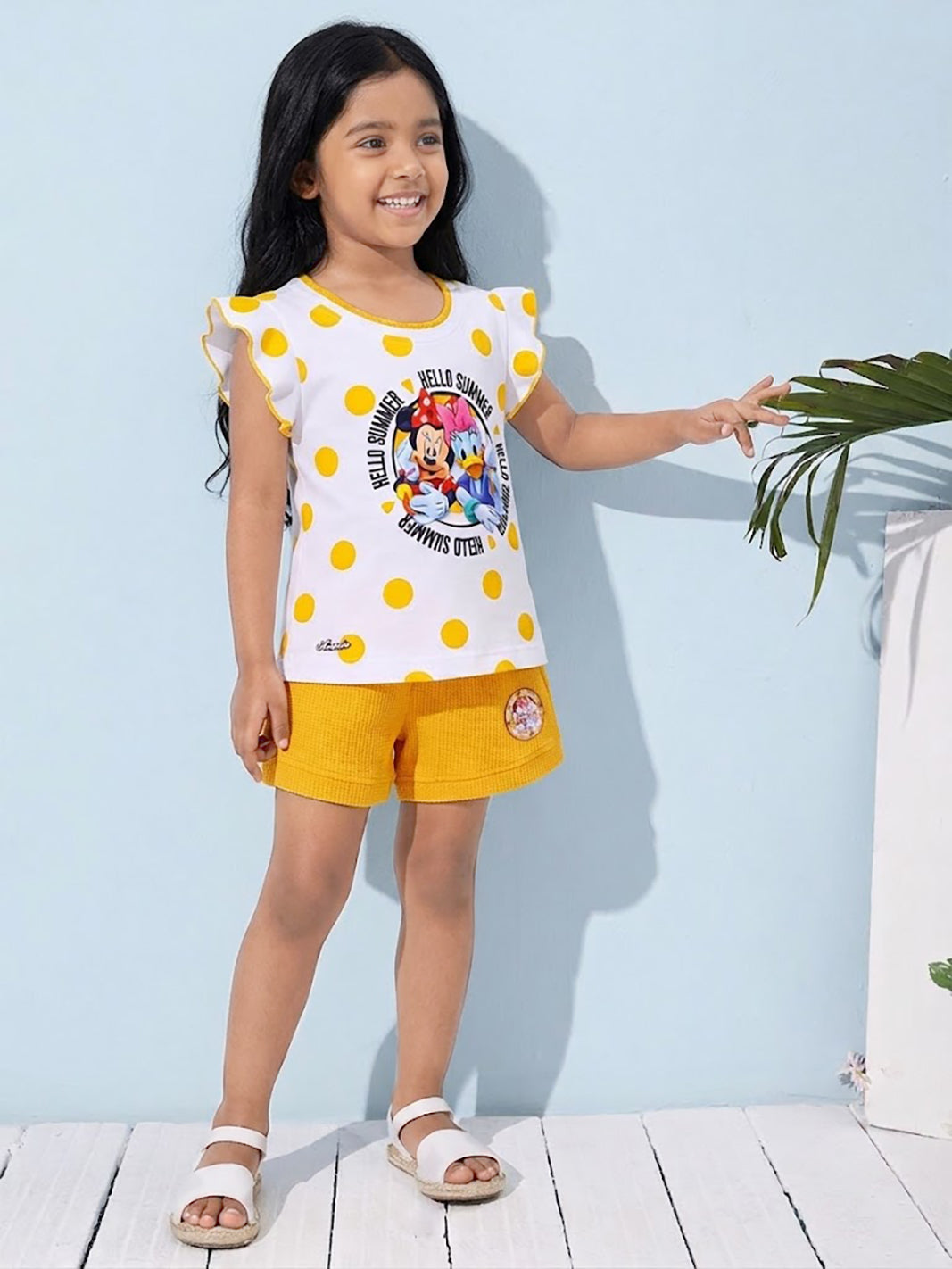 Flat lay of an Annie brand girls two-piece clothing set featuring a white flutter-sleeve top with yellow polka dots and a Minnie Mouse and Daisy Duck Hello Summer graphic paired with bright yellow textured shorts featuring belt loops and a small character patch on the left leg.