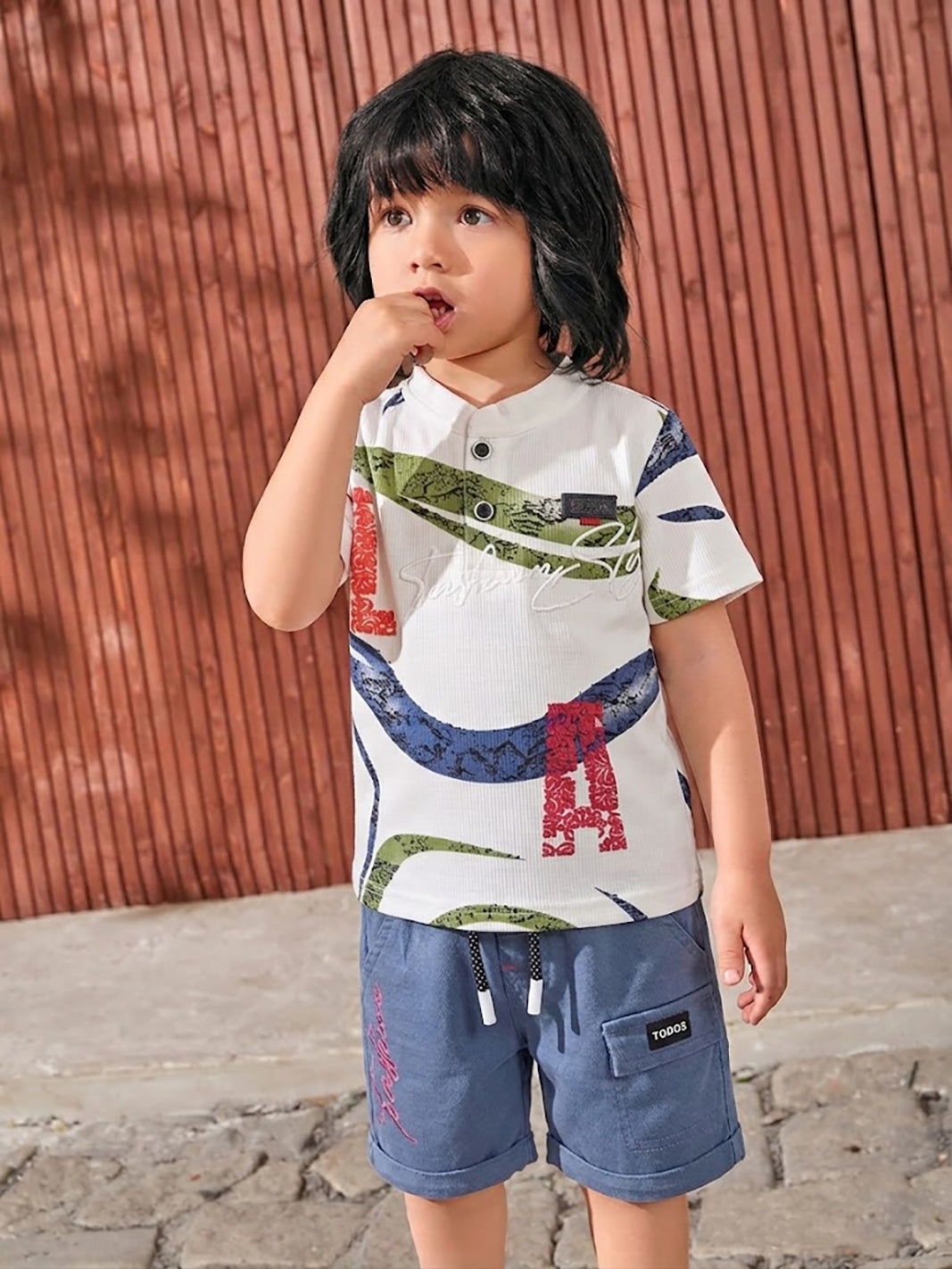 Flat lay of a TODOS brand boys two-piece clothing set featuring a white textured short-sleeve henley shirt with blue and green abstract waves and large red LA letters, paired with blue shorts with a black and white drawstring waist, Fashion embroidered in red on the right leg, and a TODOS logo patch on the left cargo pocket.