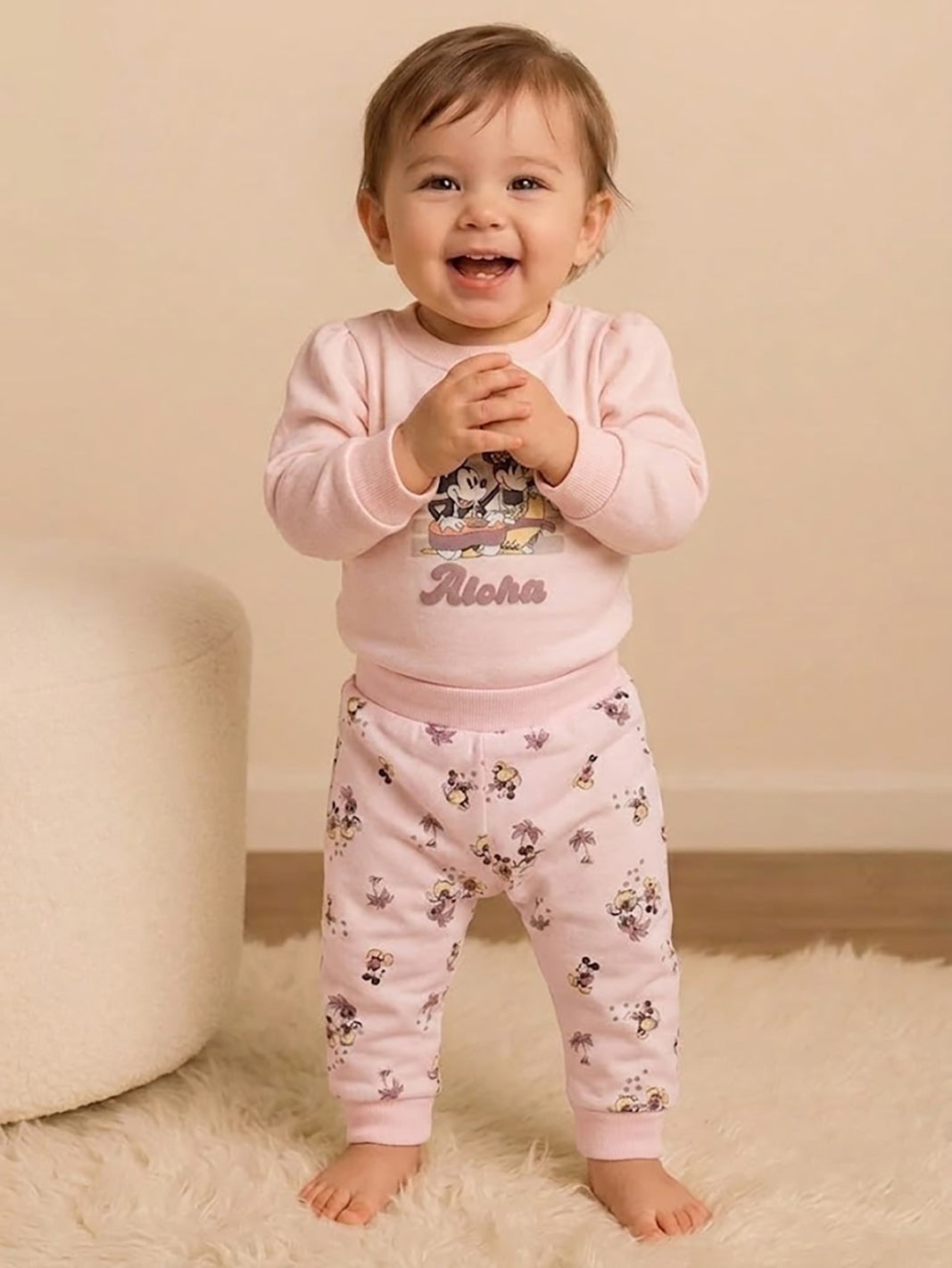 Image showing a flat lay of an Annie brand girls two-piece clothing set. The outfit includes a light pink long-sleeve top with a "GOOD TIMES Aloha" graphic featuring Mickey and Minnie Mouse, paired with matching light pink joggers covered in a whimsical all-over print of Mickey and Minnie with palm trees.