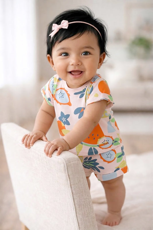 Flat lay of an Annie brand girls two-piece clothing set featuring a baby pink short-sleeve t-shirt and matching shorts with a vibrant all-over tropical fruit print including watermelon pineapple and dragon fruit. The shorts have an elastic waist with a pink decorative tie front pockets and cuffed hems.