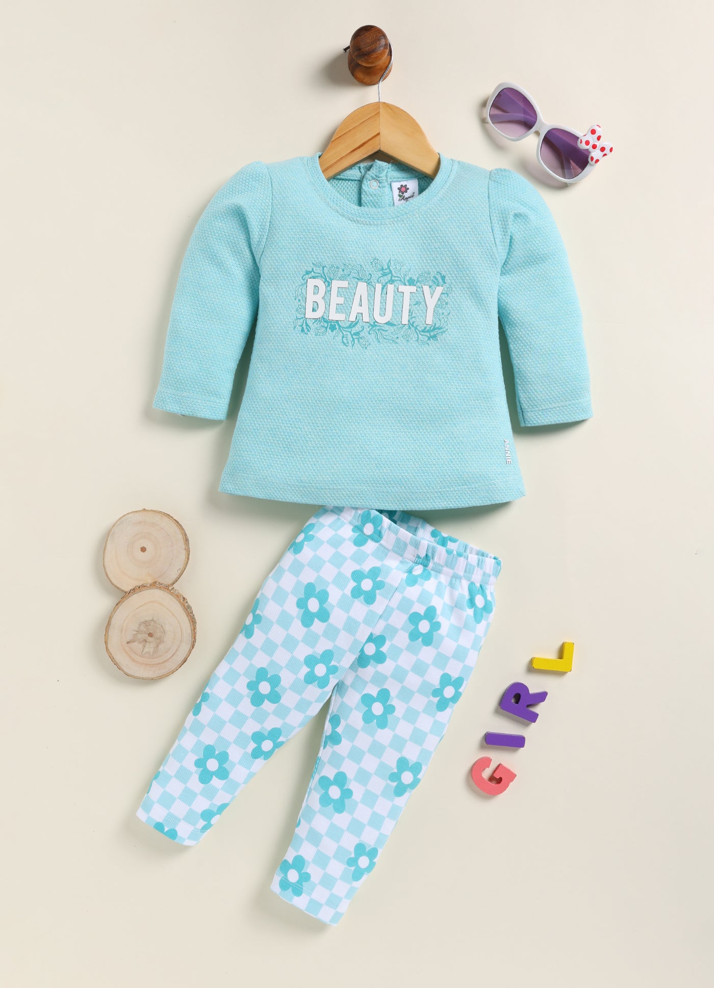 Image file BG-415-SkyBlue.jpg showing a flat lay of an Annie brand girls two-piece clothing set. The outfit includes a textured sky-blue long-sleeve top with the word "BEAUTY" and floral accents, paired with matching leggings featuring a light blue and white checkerboard pattern decorated with simple flowers.