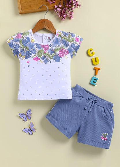 Flat lay of an Annie brand girls two-piece clothing set featuring a white short-sleeve top with a blue floral and berry print across the chest and scattered blue dots paired with textured blue shorts with an elastic tie waist white side piping and a pink cherry patch on the left leg.
