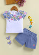 Flat lay of an Annie brand girls two-piece clothing set featuring a white short-sleeve top with a blue floral and berry print across the chest and scattered blue dots paired with textured blue shorts with an elastic tie waist white side piping and a pink cherry patch on the left leg.
