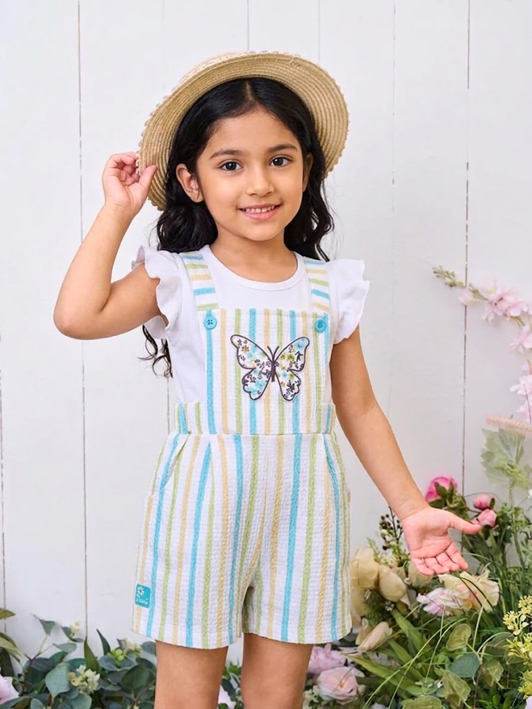 A smiling young girl modeling an Annie brand two-piece clothing set featuring a white flutter-sleeve top under textured short overalls with pastel blue, yellow, and green vertical stripes and a large floral butterfly patch on the chest.