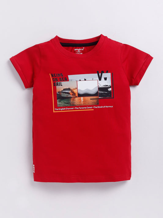 showing a young boy with curly hair modeling a Biskid brand bright red short-sleeve t-shirt. The shirt features a rectangular chest graphic with a sunset, a cruise ship, and the text "BEING ON SEA SAIL" along with famous global waterways.