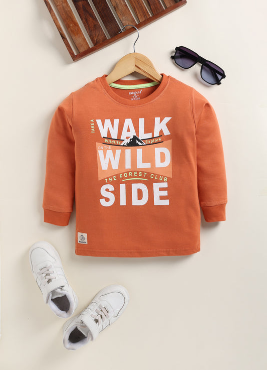 Image file Gemini_Generated_Image_pobj3hpobj3hpobj.jpg showing a young boy wearing a rust-orange Biskid brand boys long-sleeve t-shirt with a "Walk on the Wild Side" forest graphic on the chest, paired with gray checkered shorts and white sneakers.