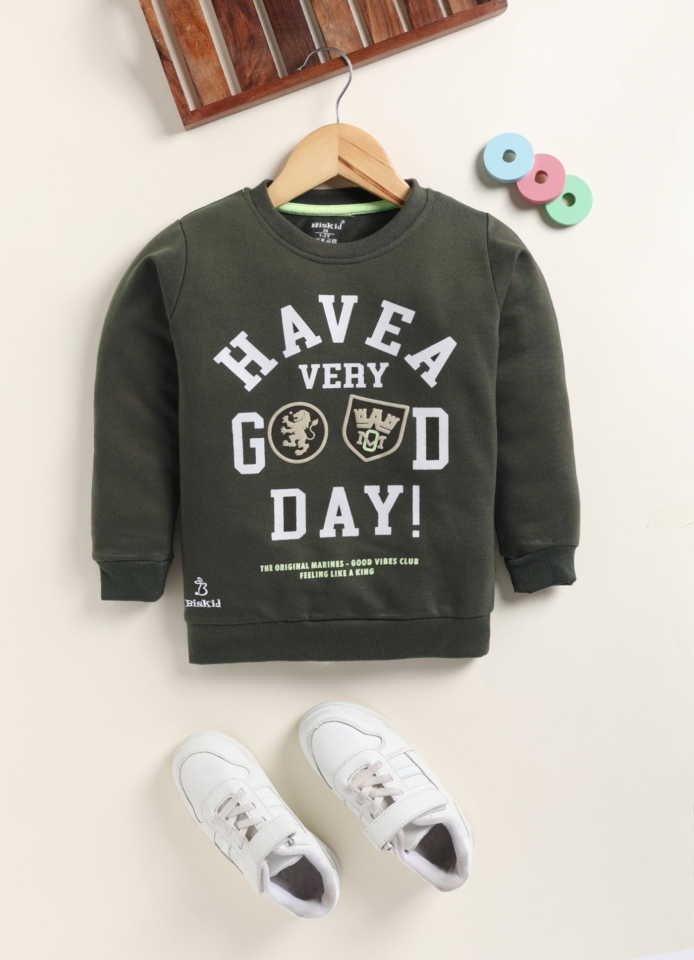 Image file Colour correction (3).jpg showing a flat lay of an olive green Biskid brand boys long-sleeve sweatshirt featuring "HAVE A VERY GOOD DAY!" text and emblem graphics, with a ribbed crew neck, cuffs, and hem.