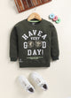 Image file Colour correction (3).jpg showing a flat lay of an olive green Biskid brand boys long-sleeve sweatshirt featuring "HAVE A VERY GOOD DAY!" text and emblem graphics, with a ribbed crew neck, cuffs, and hem.