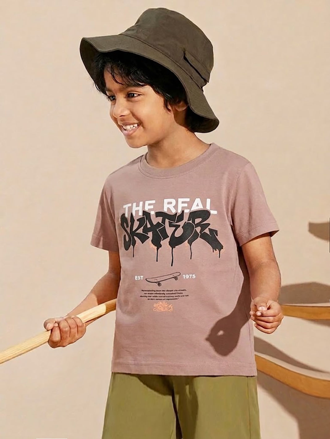 Image file 877-Lt.Brown.jpg showing a Biskid brand boys' light brown crew neck t-shirt with a bold graffiti-style "THE REAL SKATER" graphic on the front, displayed on a wooden hanger with a wooden penguin toy and a small toy airplane nearby.
