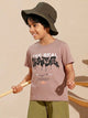 Image file 877-Lt.Brown.jpg showing a Biskid brand boys' light brown crew neck t-shirt with a bold graffiti-style "THE REAL SKATER" graphic on the front, displayed on a wooden hanger with a wooden penguin toy and a small toy airplane nearby.