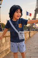 Image file 10391-Navy.jpg showing an Unokids brand boys' clothing set consisting of a navy blue graphic t-shirt with a crossbody bag print and matching light blue cargo shorts, displayed with children's white sneakers, sunglasses, and a giraffe-shaped rattle.