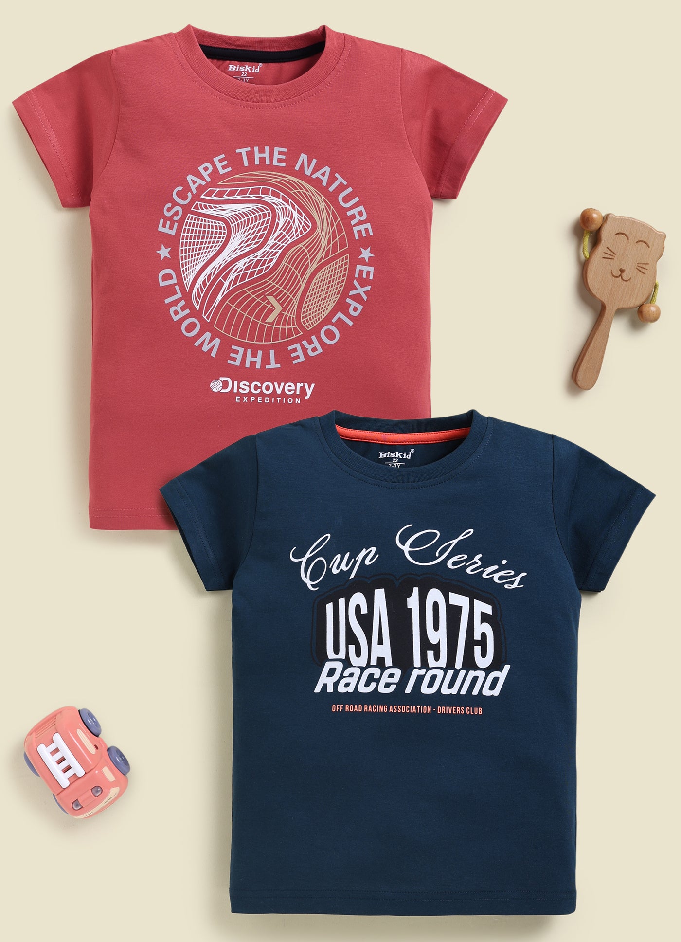 Image file 877-Blu--877-Rust.jpg showing a 2-pack of Biskid boys short-sleeve t-shirts featuring graphic prints, including a rust-red tee with "Discovery Expedition" circular graphics and a navy blue tee with "Cup Series USA 1975" typography.