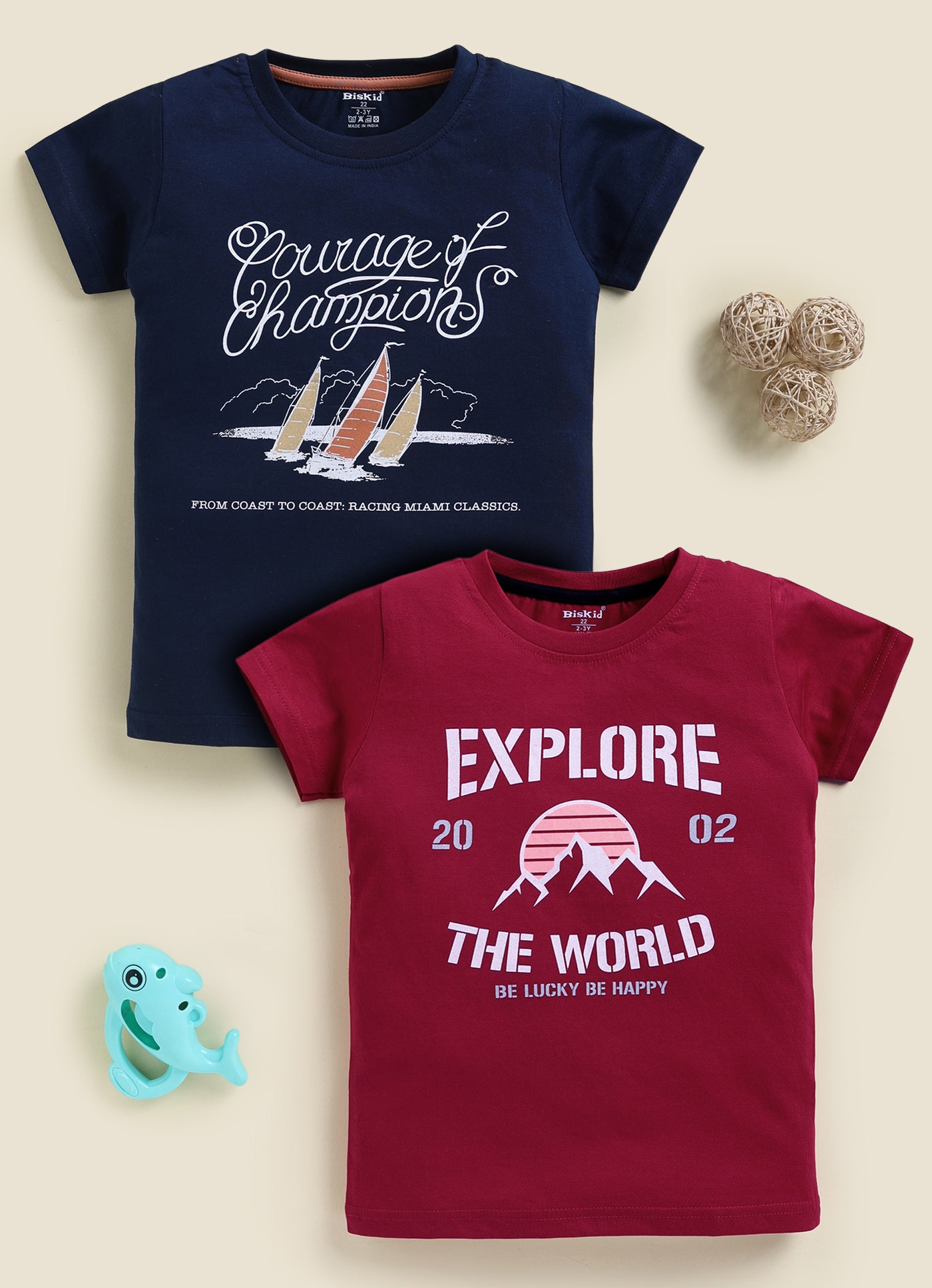 Image file 878-Nvy--878-Mron.jpg showing a 2-pack of Biskid boys short-sleeve t-shirts featuring graphic prints, including a navy blue tee with a "Courage of Champions" sailboat graphic and a maroon red tee with an "EXPLORE THE WORLD" mountain sunset print.