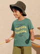 Image file 877-Green.jpg showing a Biskid brand boys' forest green crew neck t-shirt with a bold "PERFORM" graphic print on the front, displayed on a wooden hanger with small wooden spinning tops and colorful wooden "KIDS" letters nearby.