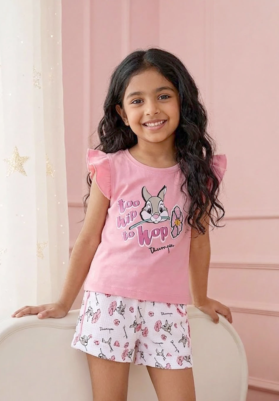 Flat lay of an Annie brand girls two-piece clothing set featuring a peach pink flutter-sleeve top with a too hip to hop Thumper rabbit graphic paired with white textured shorts featuring an all-over Thumper and pink flower print an elastic drawstring waist and pink pocket trim.