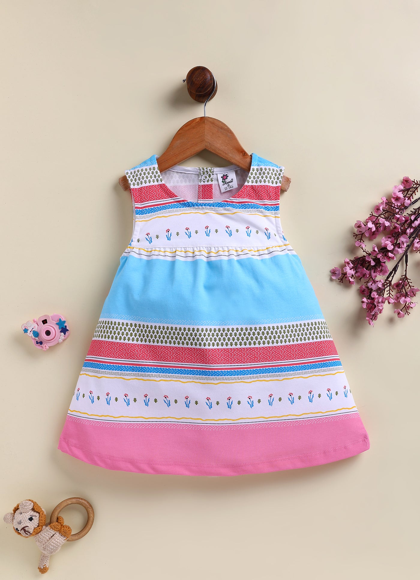 Flat lay of an Annie brand girls sleeveless frock featuring a gently gathered waist and colorful horizontal stripes in bright blue red pink and white decorated with small floral and geometric leaf patterns.