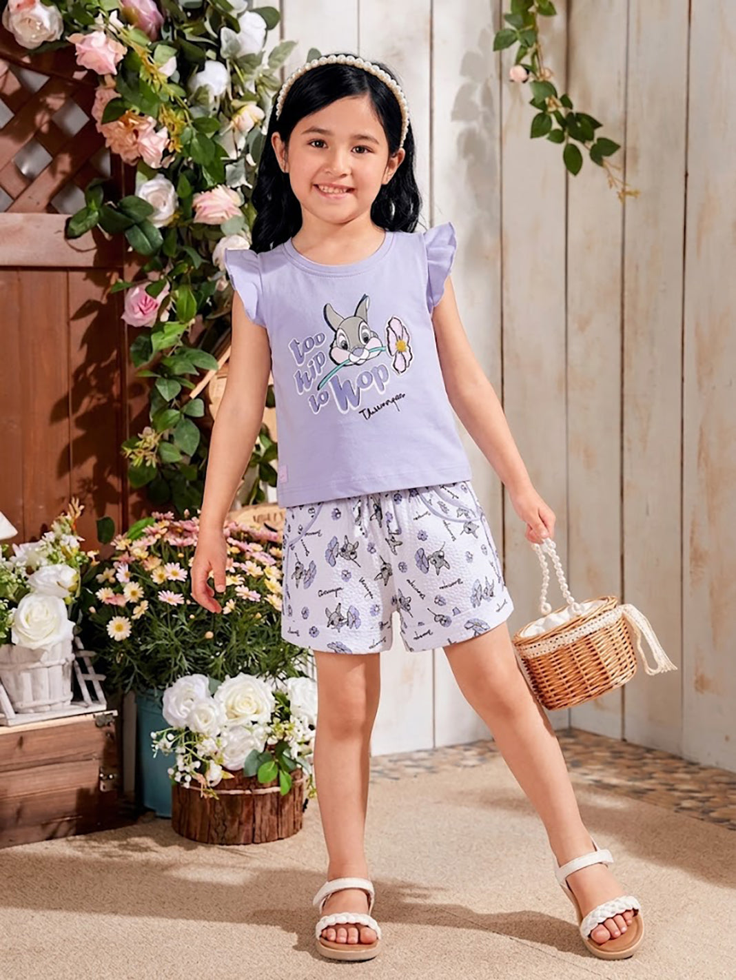 Flat lay of an Annie brand girls two-piece clothing set featuring a light purple flutter-sleeve top with a too hip to hop Thumper rabbit graphic paired with white textured shorts featuring an all-over Thumper and purple flower print an elastic drawstring waist and purple pocket trim.