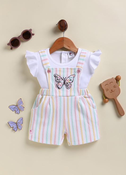 A young girl making a playful face and posing in an Annie brand two-piece clothing set featuring a white flutter-sleeve top under textured short overalls with pastel pink, yellow, and light blue vertical stripes and a large floral butterfly patch on the chest.