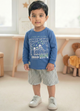 Image file Gemini_Generated_Image_u1773xu1773xu177.jpg showing a young boy wearing a blue Biskid brand boys long-sleeve t-shirt with a "Gamer" gaming controller graphic on the chest, paired with gray checkered shorts and white sneakers.