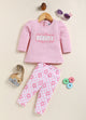 showing a flat lay of an Annie brand girls two-piece clothing set. The outfit includes a textured pink long-sleeve top with the word "BEAUTY" and floral accents, paired with matching leggings featuring a pink and white checkerboard pattern decorated with simple flowers.