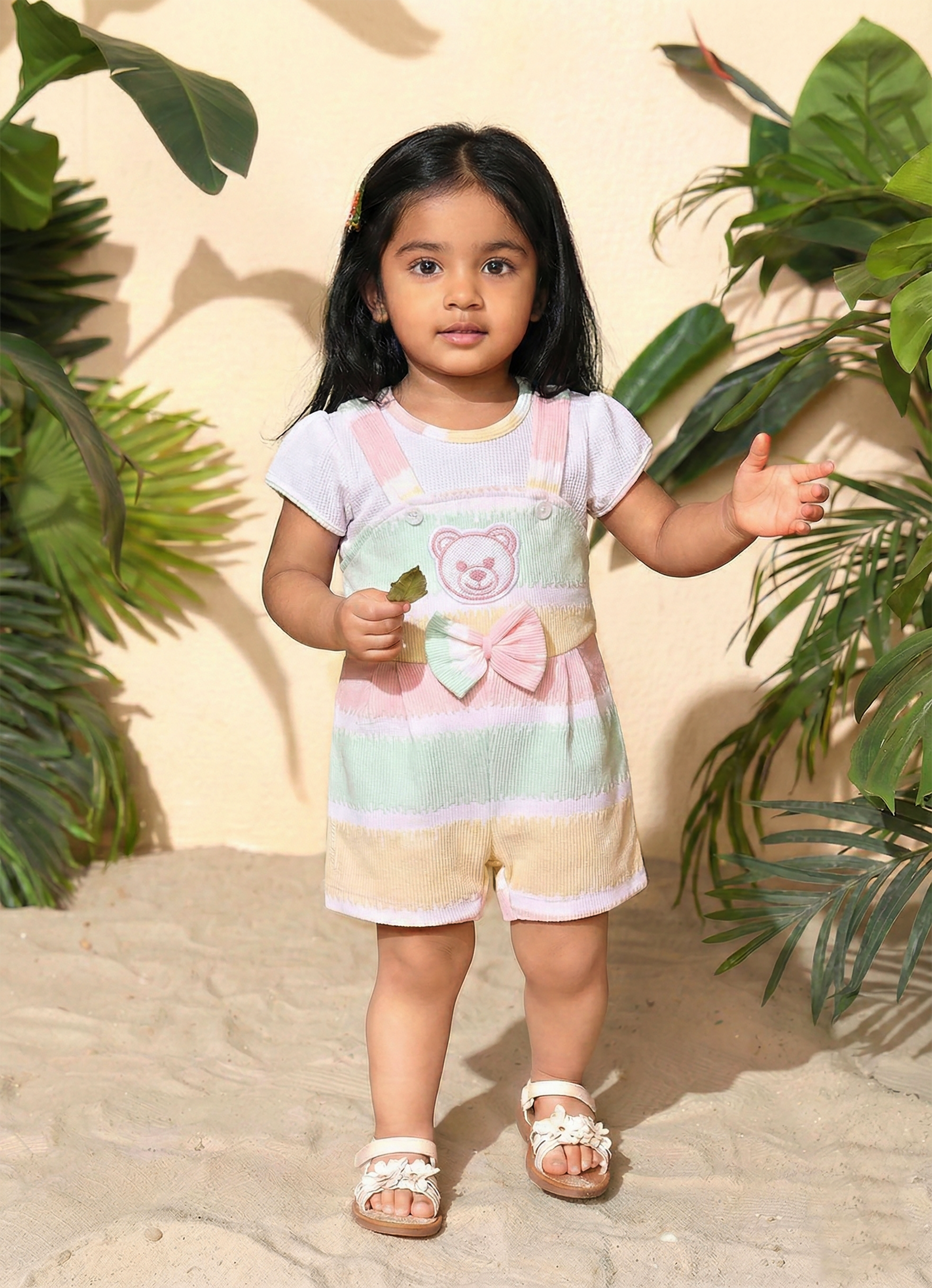 Annie brand girls short-sleeve jumpsuit designed to look like a textured white top underneath pastel green, yellow, and pink striped overall shorts. The outfit features an embroidered pink bear face on the chest, a decorative striped bow, and a back snap closure.