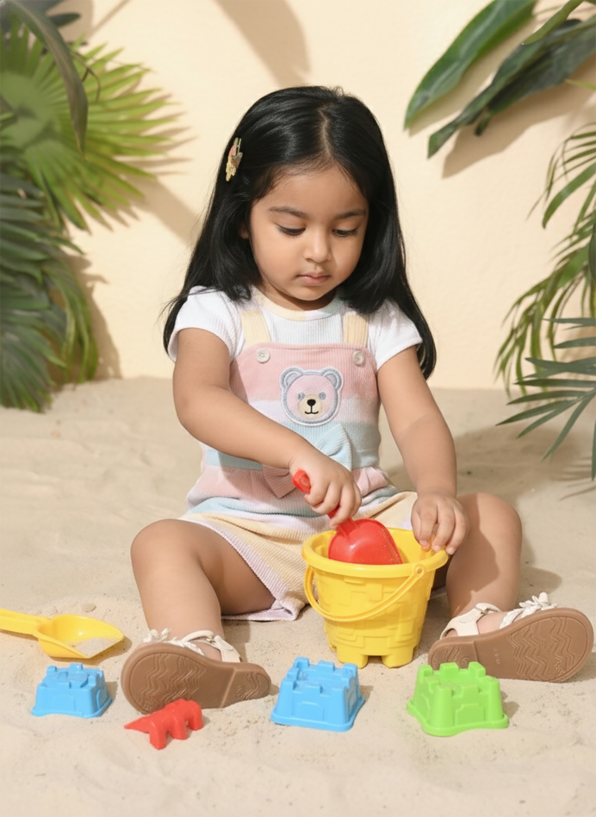  Annie brand girls short-sleeve jumpsuit designed to look like a textured white top underneath pastel pink, blue, and yellow striped overall shorts. The outfit features an embroidered bear face on the chest, a decorative bow, and a back snap closure.