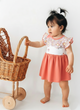 Annie brand girls two-piece frock and panty set. The dress features a white flutter-sleeve bodice with a coral red daisy and butterfly print, a back snap closure, and a solid coral red gathered skirt. It comes with a matching white bloomer panty featuring coral red ruffled trim on the leg openings.