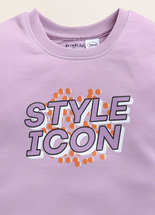 Image file 1754-Purple.jpg showing a Biskid brand girls lavender-purple sweatshirt featuring the text "STYLE ICON" on the front, displayed on a wooden hanger with children's boots and a heart-shaped woven decoration nearby.