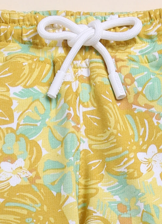 Image file 1511-Mustard.jpg showing a pair of Biskid brand girls mustard yellow joggers with an all-over tropical leaf print, displayed with fun decorative accents like sunglasses and wooden "CUTE" letters.
