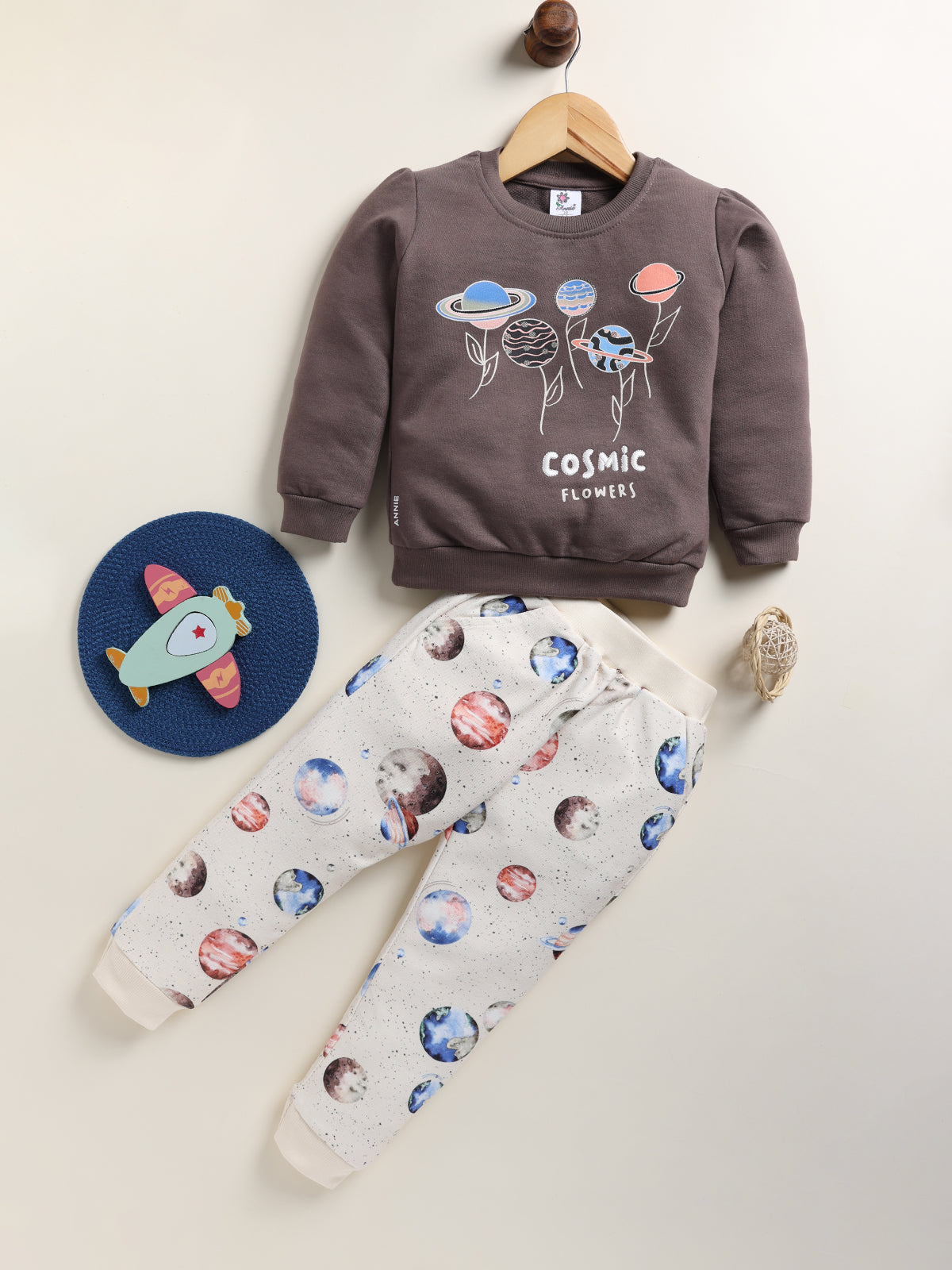 Image showing a flat lay of an Annie brand girls two-piece clothing set. The outfit includes a sage long-sleeve sweatshirt with a "COSMIC FLOWERS" graphic featuring stylized planets, paired with grey joggers covered in an all-over whimsical print of various planets and stars.