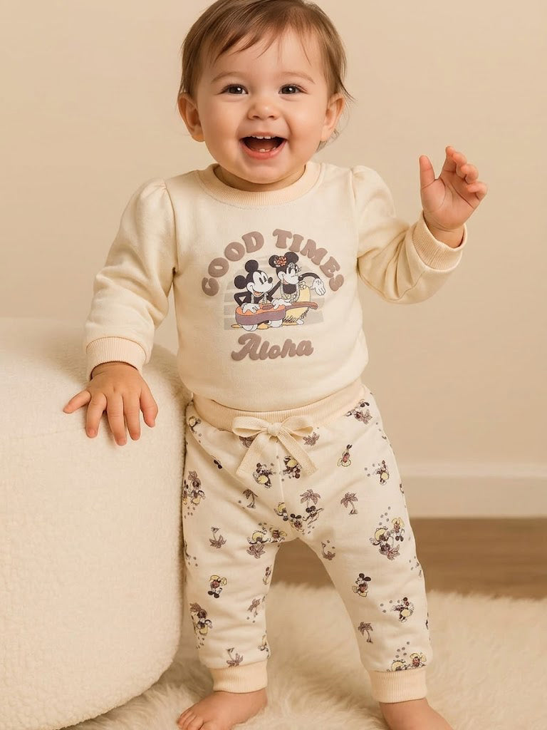 Image showing a flat lay of an Annie brand girls two-piece clothing set. The outfit includes a cream-colored long-sleeve top with a "GOOD TIMES Aloha" graphic featuring Mickey and Minnie Mouse, paired with matching cream-colored joggers covered in a whimsical all-over print of Mickey and Minnie with palm trees.