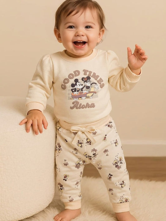 Image showing a flat lay of an Annie brand girls two-piece clothing set. The outfit includes a cream-colored long-sleeve top with a "GOOD TIMES Aloha" graphic featuring Mickey and Minnie Mouse, paired with matching cream-colored joggers covered in a whimsical all-over print of Mickey and Minnie with palm trees.
